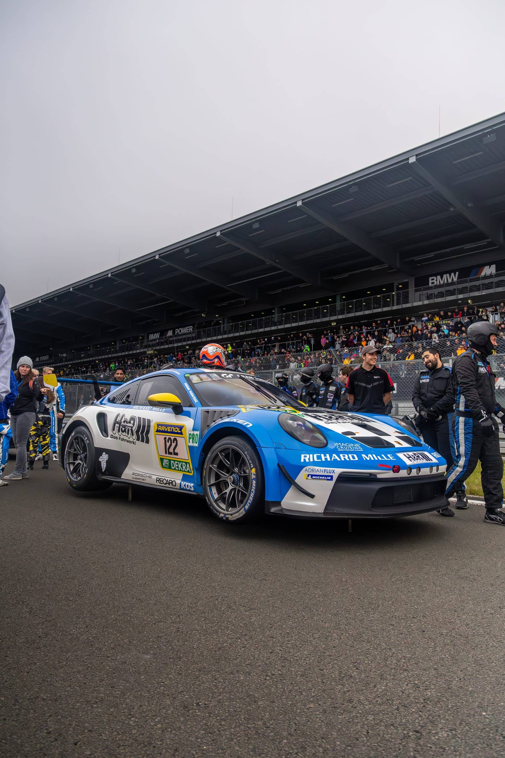 A blue and white race car staged at trackside with grandstands in the background.