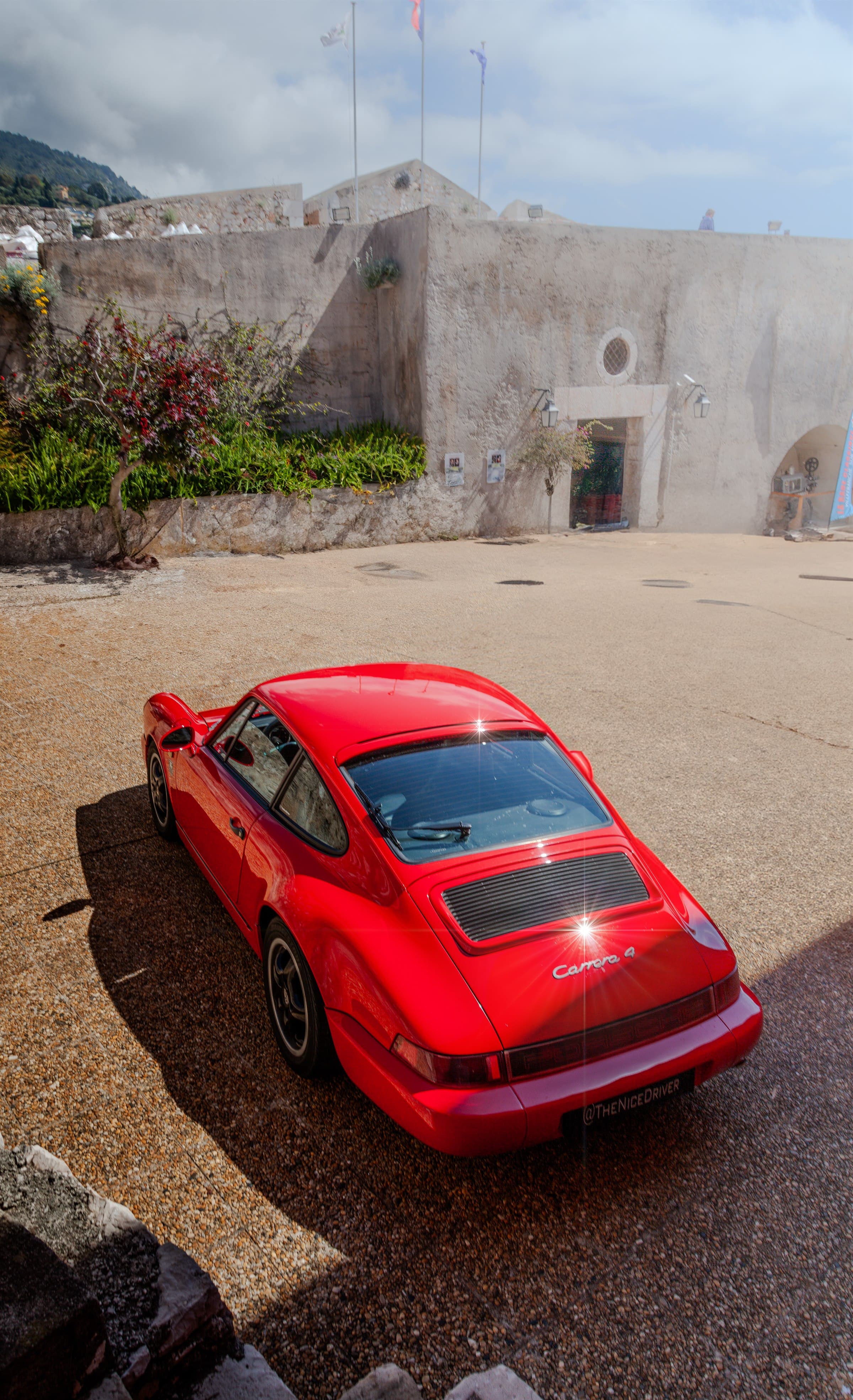 A red coupe framed on a stone road beside a textured architectural wall.