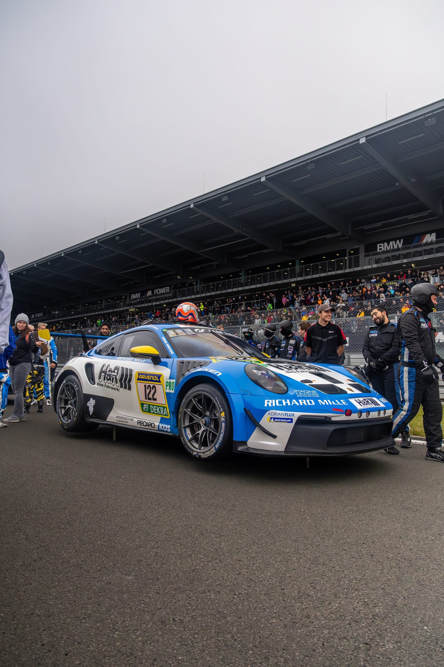 Blue and white GT car on the grid among photographers and crew.