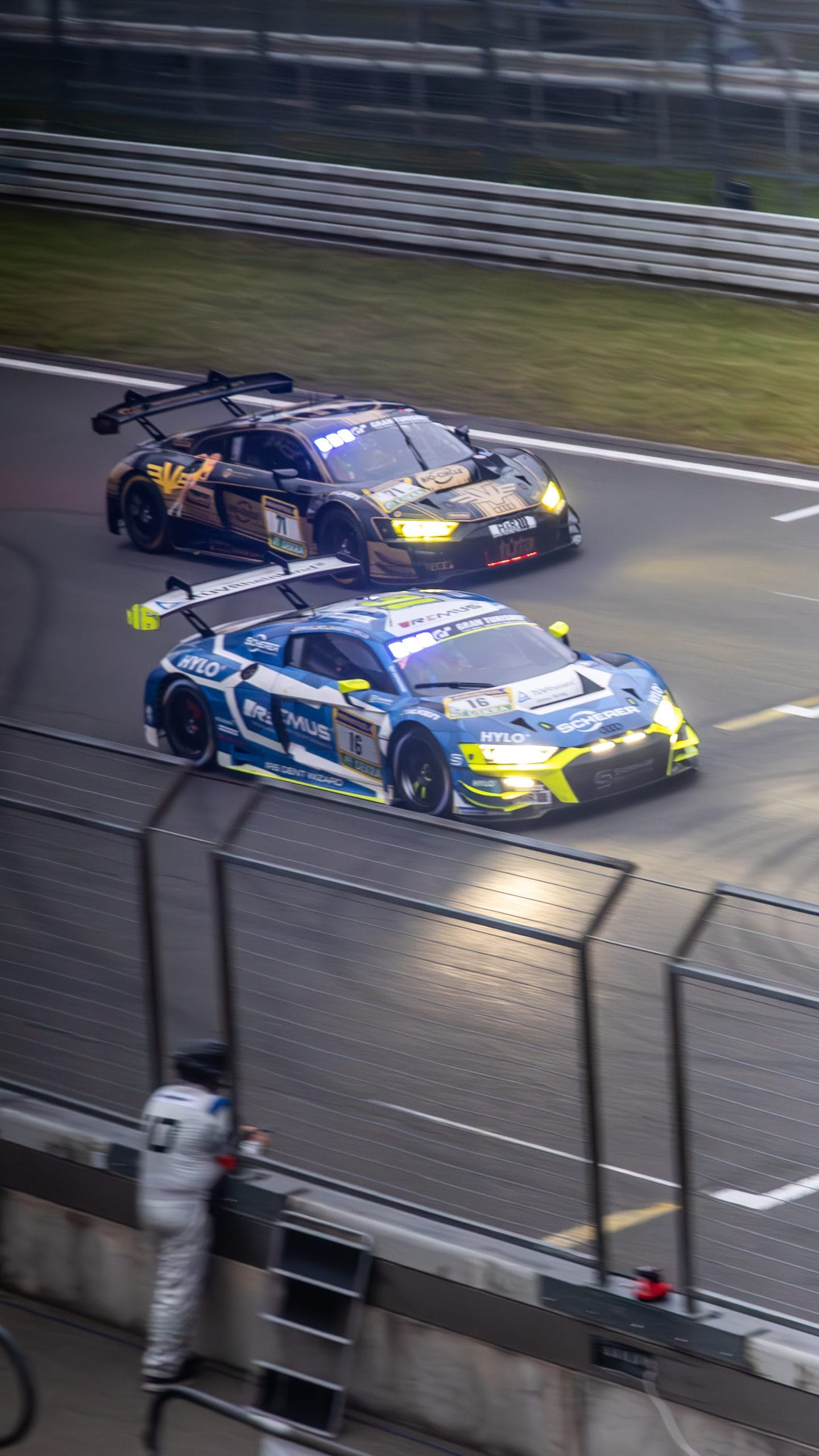 Two blue racing cars on track in the evening, a marshal at the fence.