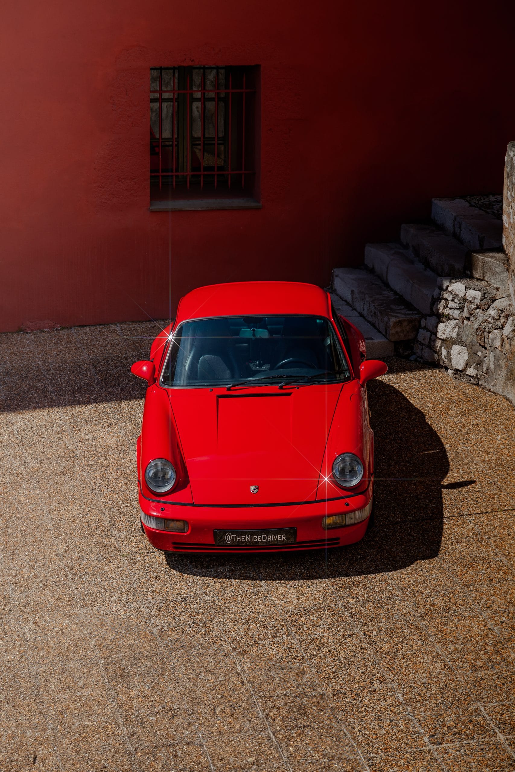 Red Porsche 911 seen from above in a shaded courtyard with a red wall.