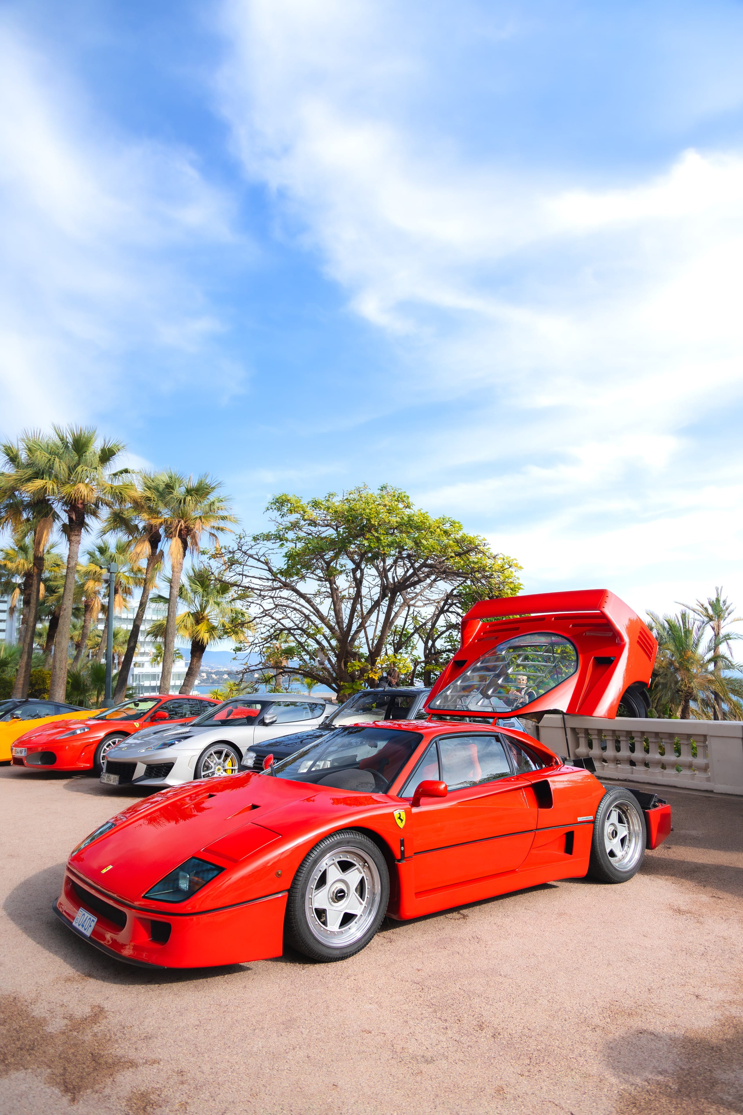 Red supercar with its rear clamshell raised, on a terrace above the sea, palms behind.
