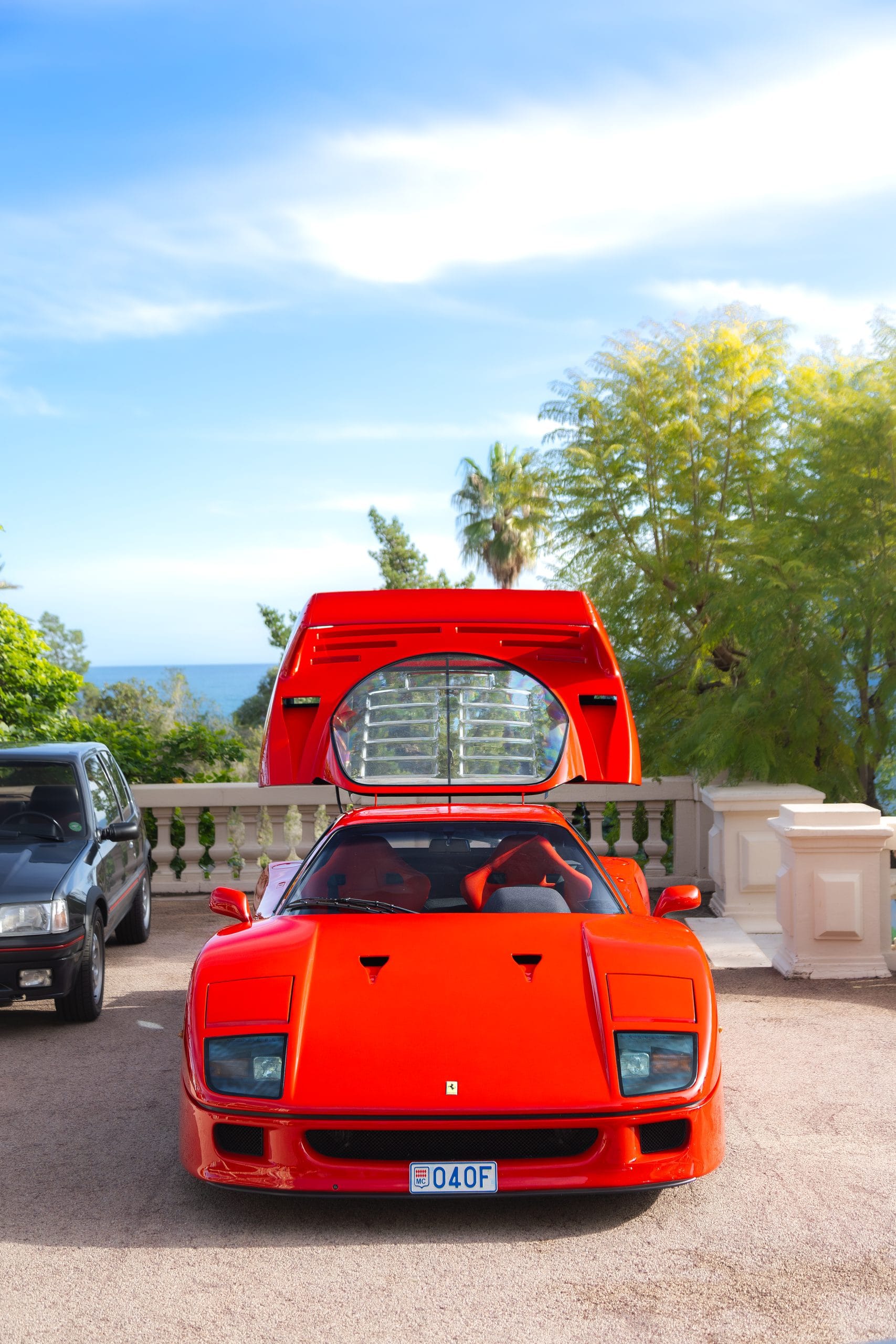 The red supercar from behind, rear clamshell open, on a sunlit terrace.