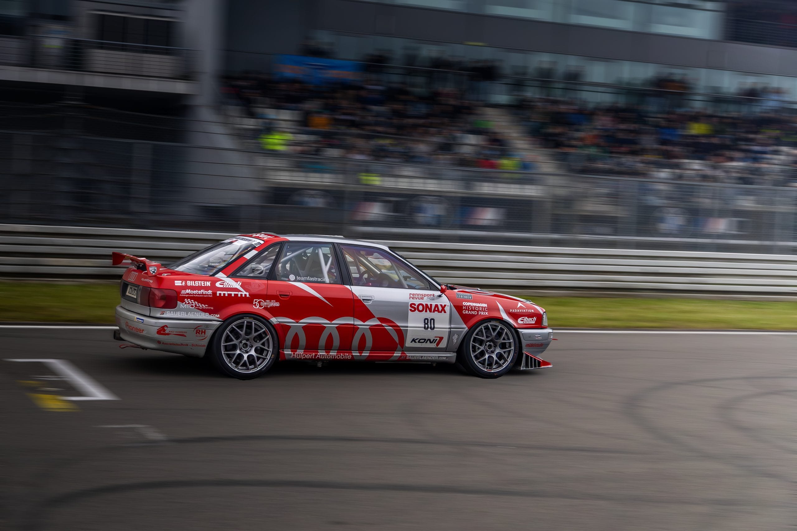 A red and white touring car at speed, the grandstand blurred behind it.