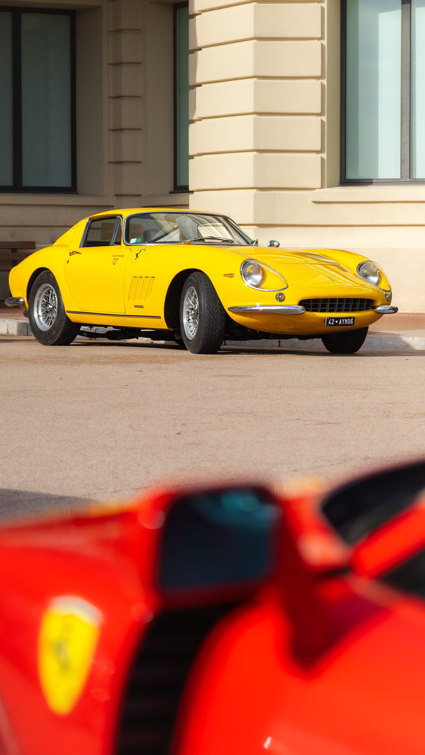 Yellow classic coupe framed by a blurred red bodywork in the foreground.