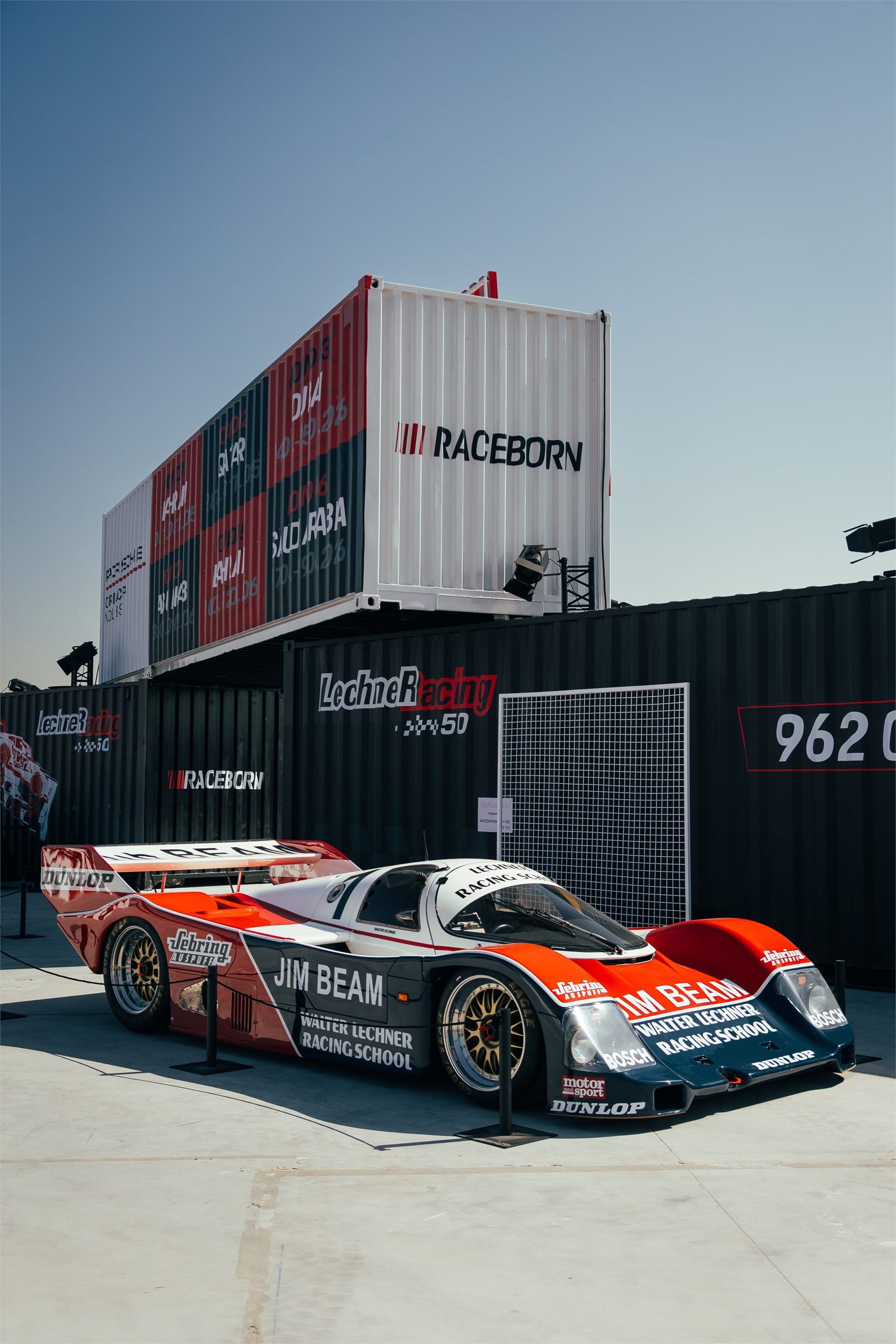 White and red Le Mans racing car in front of stacked shipping containers.