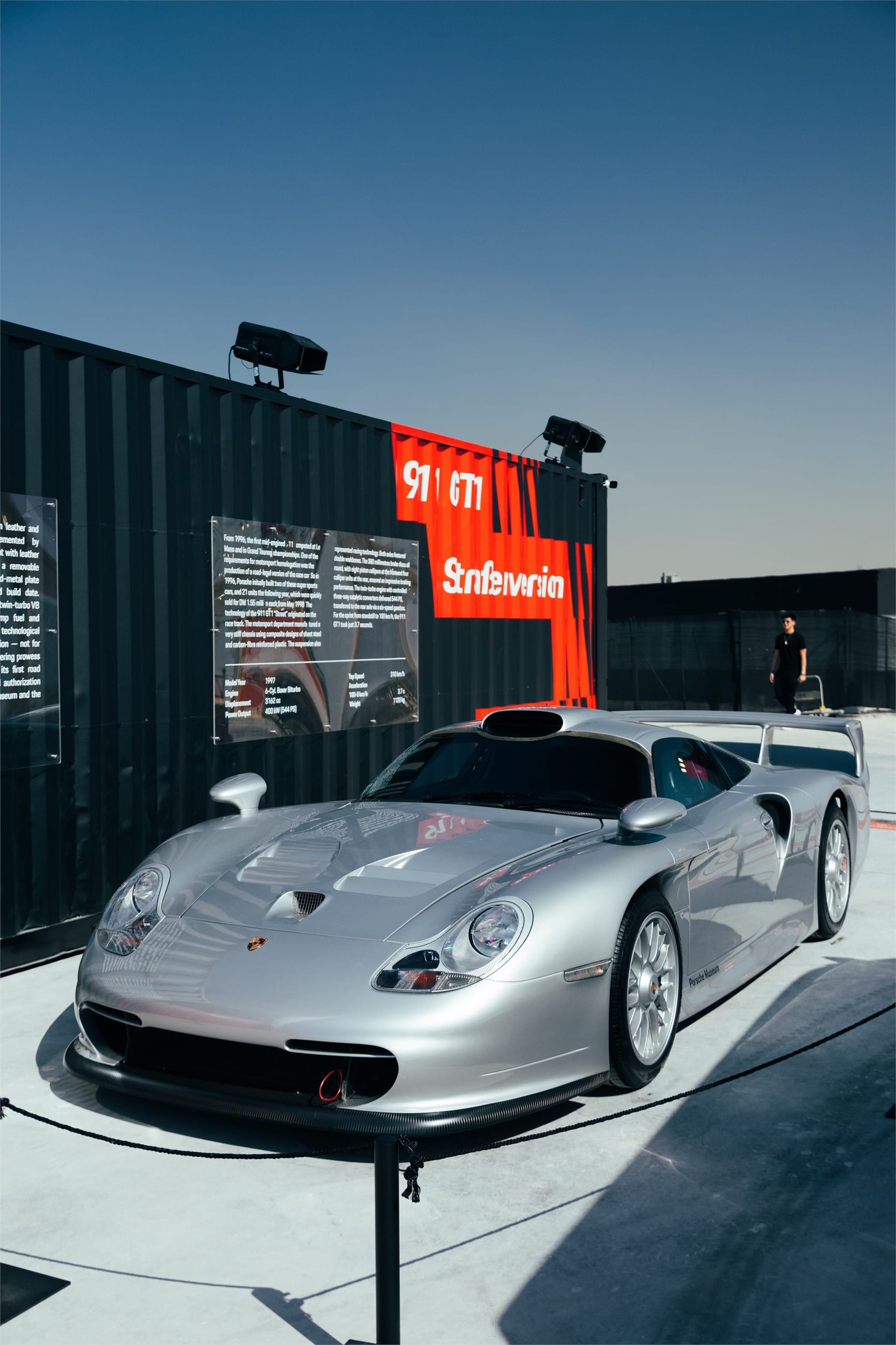 Silver GT racing car on display beside a black container.