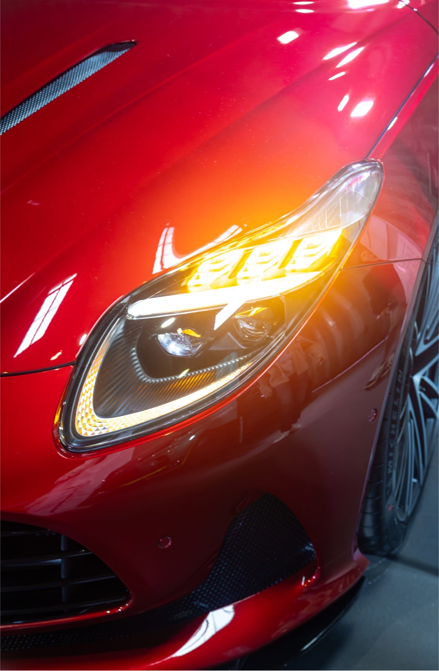 Lit headlight of a red Aston Martin.